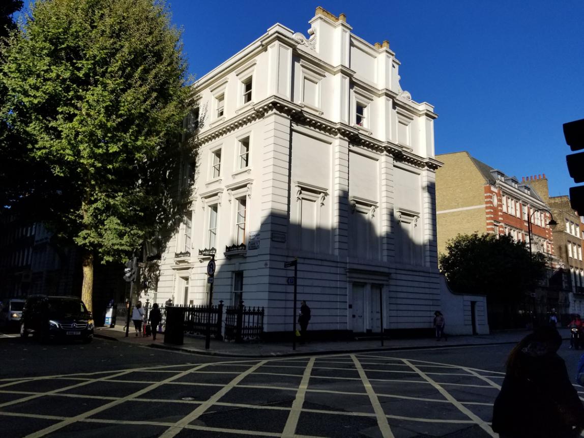 A multi-story white 18th-century Georgian building stands at the intersection of two streets. A large tree stands to the left of the building.
