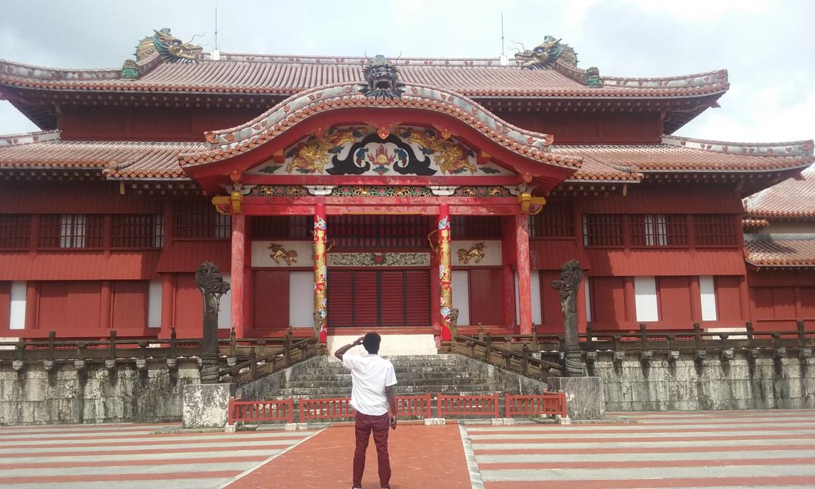 A student stands in awe of a large building in Okinawa. The building is multi-tiered and bright red, with ornate detail work above and around the front door.