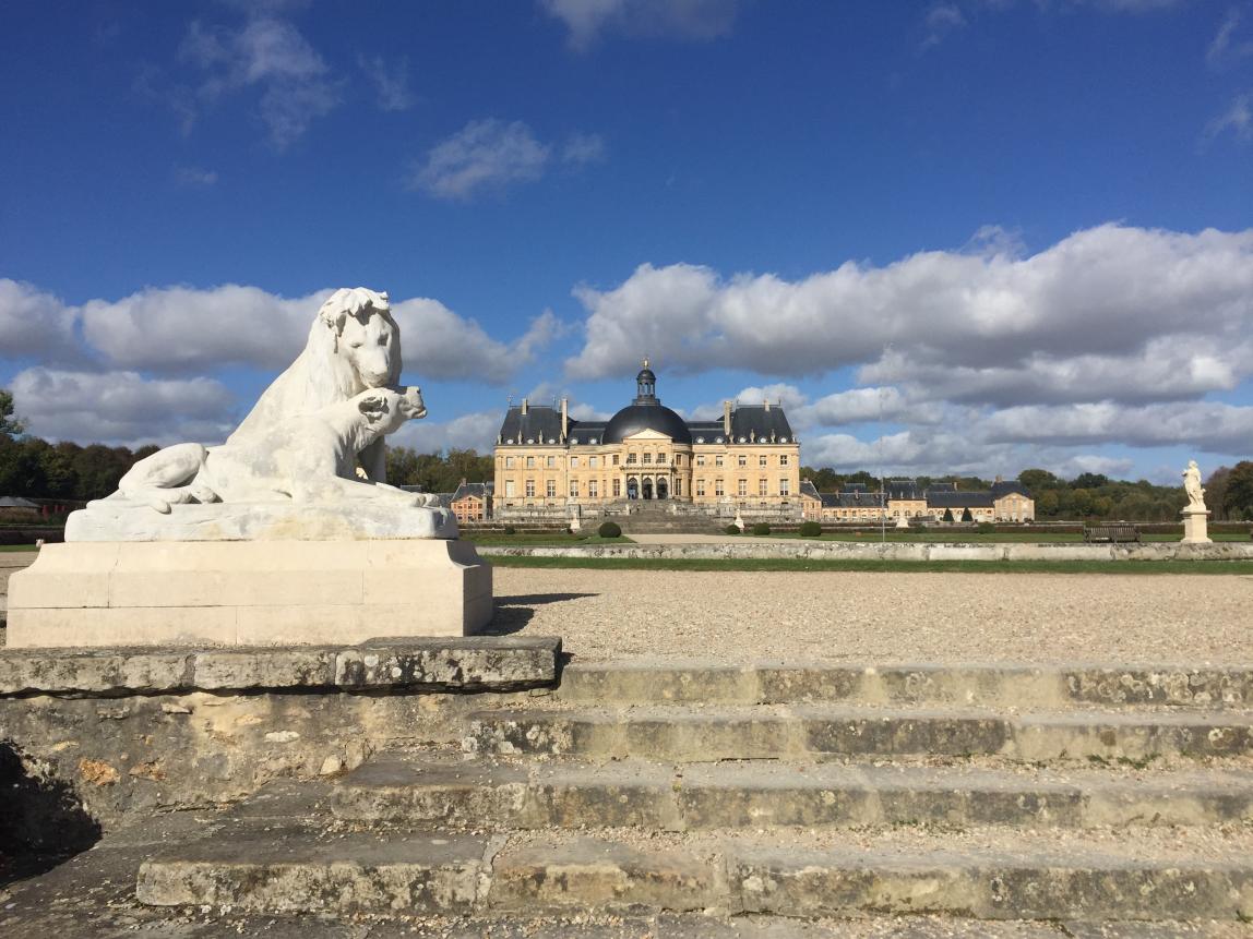 A statue of two lions sitting together in the foreground of the palace of Fontainebleau. A building with a cream exterior and dark roof, along with other statues, sit in the background.