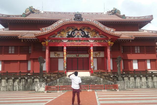 A student stands in awe of a large building in Okinawa. The building is multi-tiered and bright red, with ornate detail work above and around the front door.