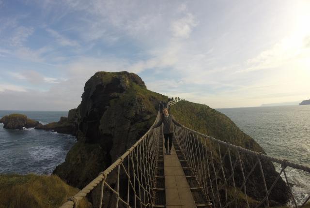 A student walks across a narrow swinging bridge over water.