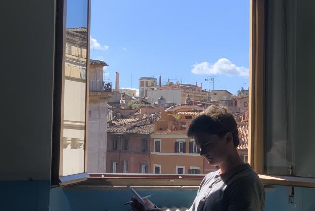 A student at the Rome Center works on their assignment with the window open. Outside the window are terracotta-colored buildings.