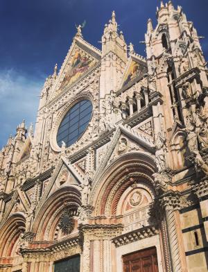 A cathedral in Siena, the Duomo Di Siena, on a sunny day. The exterior is an off-white color, with ornate details on the structure.