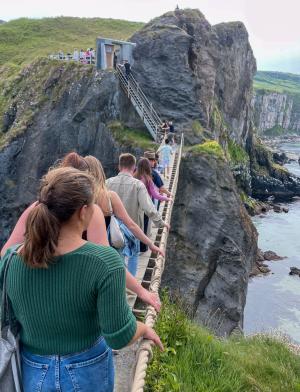 line of students walking on a rope bridge across an oceanside mountanous terrain